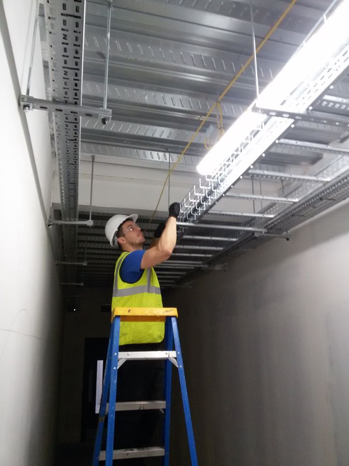 An electrician in a hi-vis vest and hard hat on a stepladder, fitting cable tray and strip lighting along a corridor ceiling