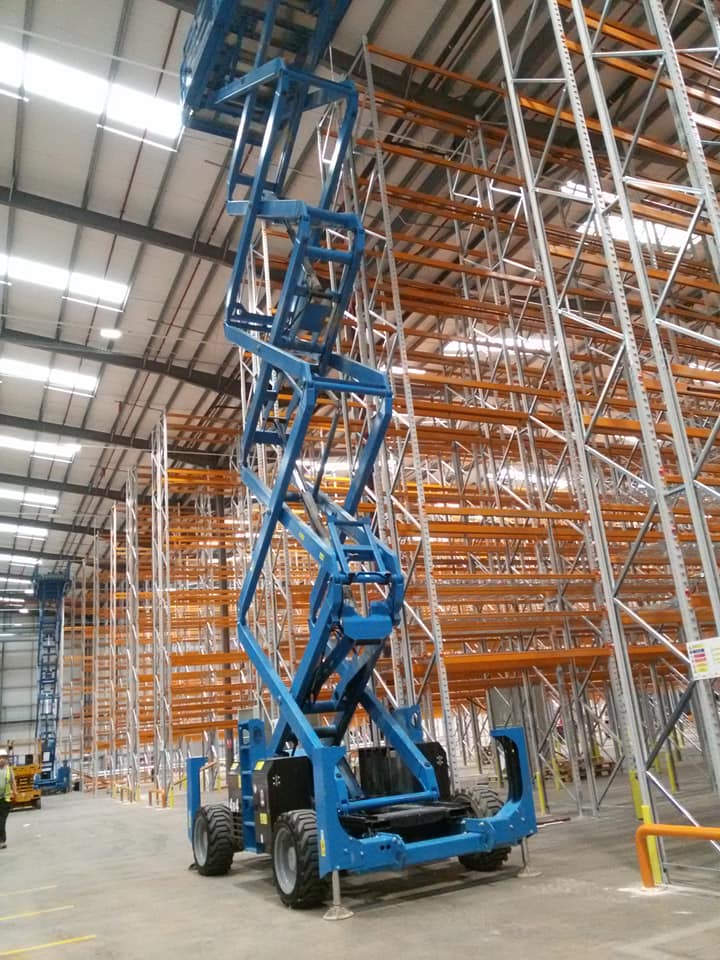 A blue access platform raised between orange pallet racking in a large warehouse