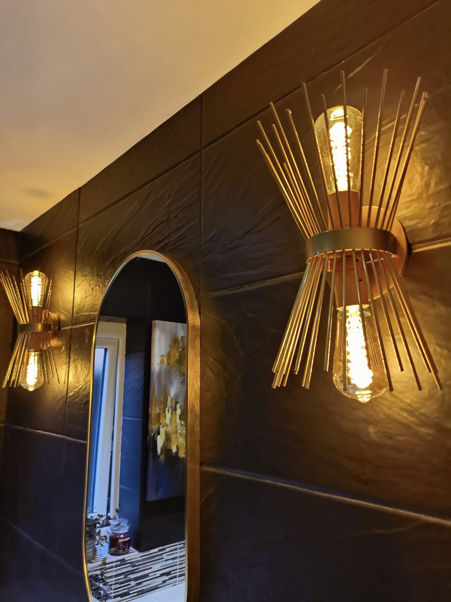 Close view of two gold sunburst wall lights on a dark tiled bathroom wall beside an oval mirror